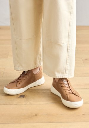 Hnědé kožené tenisky s perforovanými detaily, texturované bílé gumové podrážky a tradiční šněrování, nošené s béžovými kalhotami se širokými nohavicemi.