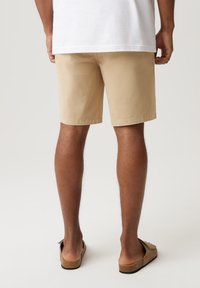 Beżowe bawełniane szorty o prostym kroju, z podwiniętymi brzegami i luźnym fasonem, w połączeniu z brązowymi klapkami. Widok z tyłu.