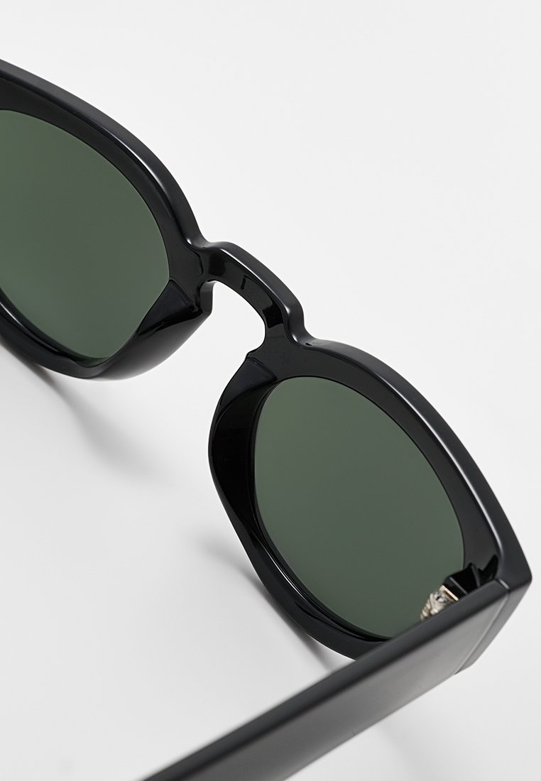 Lunettes noires avec des verres verts ronds, une monture épaisse et une légère courbure au niveau du pont, présentant une finition brillante et des branches robustes.