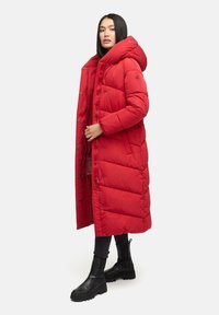 Manteau matelassé rouge avec capuche, présentant des sections matelassées, des boutons-pression et deux poches latérales. Porté sur un pantalon noir et des bottes noires imposantes.
