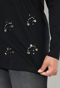 Zwarte top met lange mouwen, voorzien van metalen pailletten in decoratieve patronen. De stof lijkt glad te zijn, met een iets ontspannen pasvorm aan de zoom.