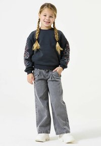 Felpa grigia scura con ricami floreali sulle maniche, abbinata a pantaloni in denim grigi chiari e scarpe da ginnastica bianche. Il bambino sorride.