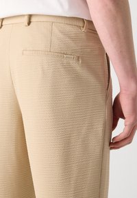Beige teksturerte bukser med et vevd mønster, sidelommer og en baklomme med et merke. Glatt overflate og skreddersydd passform.