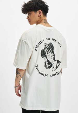 Nuori mies, jolla on tatuointeja, pukeutunut valkoiseen ylisuurikokoiseen t-paitaan, jossa on rukoilevat kädet pitämässä rahaa ja teksti "Money on my mind".