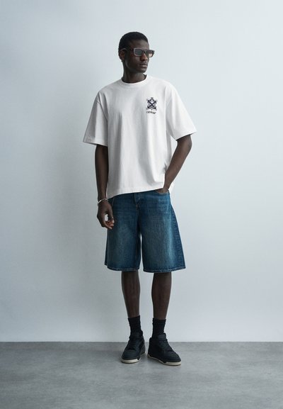 Uomo con maglietta grafica bianca oversize, pantaloncini di jeans fino al ginocchio, calzini neri, sneakers nere e occhiali da sole, in piedi contro un muro bianco.