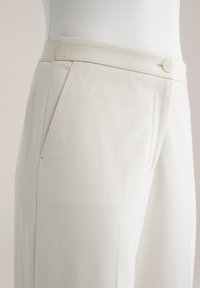 Biele nohavice na mieru s hladkou tkaninou, bočnými záhybmi a zapínaním na gombík v páse. Jednoduchý, moderný dizajn.