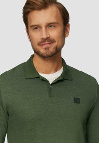 Donkergroene polo-shirt van een gestructureerde stof, met korte mouwen en een subtiele logo-patch op de linkerborst, gelaagd over een wit T-shirt.