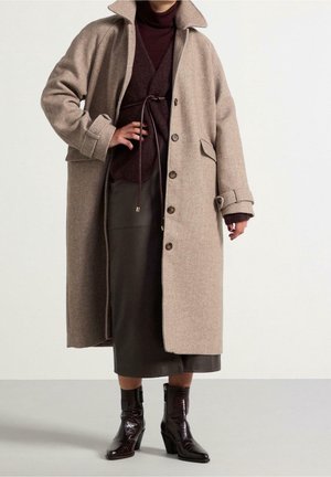 Persona con un abrigo largo beige sobre un conjunto marrón oscuro con cinturón y botas negras de cuero hasta la mitad de la pantorrilla, de pie sobre un fondo claro y liso.