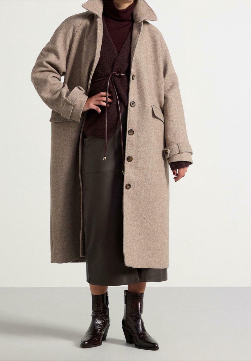 Personne portant un long manteau beige par-dessus une tenue marron foncé ceinturée et des bottes en cuir noir mi-mollet, debout sur un fond clair uni.