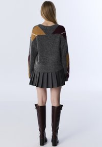 Grau gestrickter Pullover mit bunten geometrischen Mustern auf den Schultern, kombiniert mit einem grauen Faltenrock und braunen kniehohen Stiefeln.