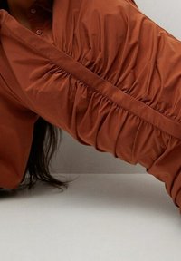 Blouse in roestkleur met lange, geplooide mouwen en een gladde textuur. Voorzien van een knoopsluiting aan de voorkant en een losse, vloeiende pasvorm.