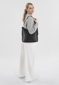 Black leather shoulder bag with a textured finish, featuring an adjustable strap and a top zipper closure. Model wearing a grey sweater and white pants.