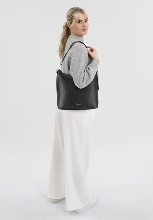 Black leather shoulder bag with a textured finish, featuring an adjustable strap and a top zipper closure. Model wearing a grey sweater and white pants.