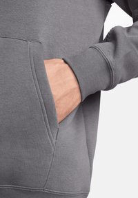 Felpa grigia con tasca a marsupio. Presenta polsini e orlo a costine, una texture liscia e cuciture del tessuto visibili attorno alla tasca.