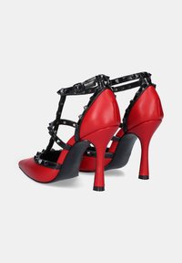 Zapatos de tacón alto rojos con puntera afilada. Incluyen correas negras con tachuelas y un tacón negro contrastante. Textura de cuero suave.