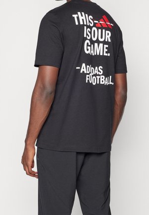 Osoba nosící černé tričko s bílým nápisem "This is our game. - Adidas Football" a červeným logem Adidas na zádech.