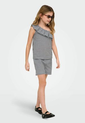 Dziewczyna ubrana w czarno-białą bluzkę w kratkę gingham z falbaną na jedno ramię oraz pasujące do niej szorty, czarne buty Mary Jane i okulary przeciwsłoneczne w oprawkach w kolorze żółwia, idzie.