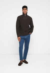 Brązowy sweter o fakturze z wysokim kołnierzem i półzamkiem, zestawiony z niebieskimi dżinsami i ciemnobrązowymi mokasynami. Prosty, nowoczesny design.