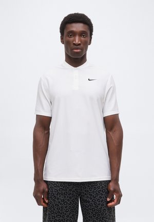 Uomo che indossa una polo Nike bianca a maniche corte e pantaloncini scuri con stampa leopardata, in piedi contro uno sfondo chiaro e uniforme.