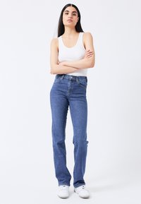 Jeans de mezclilla azul de tiro alto con corte recto, cierre de botones y bolsillos delanteros. Llevados con una camiseta de tirantes blanca con canalé y zapatillas blancas.