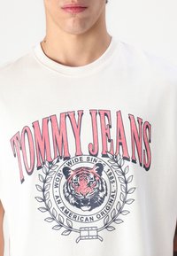 Off-white ærmeløs t-shirt med et grafisk print af en tiger omgivet af laurbærblade, med "TOMMY JEANS" i fed rød og sort tekst.
