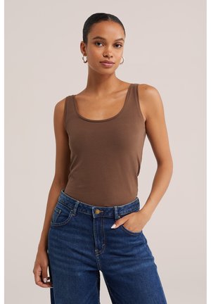 Femme avec des boucles d'oreilles créoles portant un haut marron sans manches et un jean bleu, une main dans la poche, debout devant un fond beige uni.