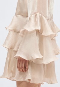 Hand rustend naast gelaagde, geplooide, lichtbeige satijnen jurkstof met een subtiele glans en een vloeiende textuur.