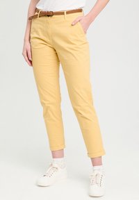 Pantalon en coton jaune à coupe slim, avec des chevilles retroussées, deux poches avant et une ceinture tressée marron. Associé à des baskets blanches et des chaussettes à motifs.