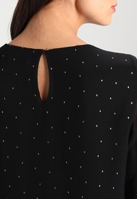 Blusa nera con apertura a chiave sul retro, caratterizzata da piccoli punti metallici color oro rosa distribuiti uniformemente sulla stoffa. Tessuto leggero e liscio.