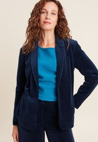 Blazer in velluto a coste blu navy con rever a ragdine, abbinato a un top aderente color teal. Il blazer presenta una texture liscia e tasche laterali.