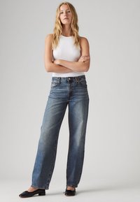 Světle modré džíny s vysokým pasem a širokými nohavicemi z denimu, s předními kapsami a klasickým zapínáním na knoflík, kombinované s bílým tílkem a černými balerínkami.