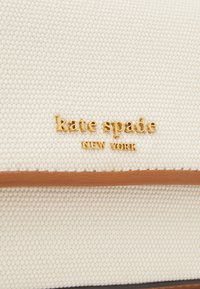Beżowa torebka z tkaniny o splocie, z złotym logo „kate spade”, brązowymi skórzanymi wykończeniami, gładką fakturą i usztywnionym kształtem.