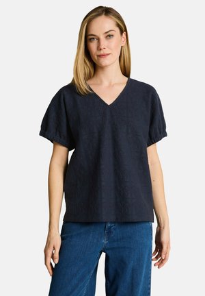 Frau mit glatten blonden Haaren, die ein dunkelblaues, strukturiertes V-Ausschnitt-Kurzarmshirt und blaue Jeans vor einem einfarbigen Hintergrund trägt.