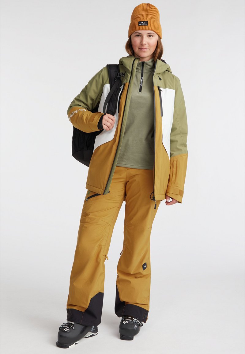 Ski-Outfit mit einer wasserdichten Jacke in Grün und Braun mit weißen Akzenten, passenden braunen Hosen, einer gestrickten orangen Mütze und schwarzen Stiefeln.