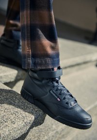 Baskets montantes en cuir noir, avec une fermeture à lacets à l'avant et une sangle à Velcro. Présentent des détails texturés et un logo de marque visible.