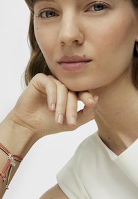 Primo piano del viso di una persona con pelle chiara, capelli lisci e un indumento bianco. Un bracciale a più fili presenta elementi rossi, marroni e argentati.
