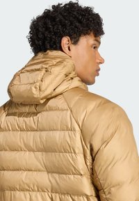 Svijetlosmeđi puffer jakna s kapuljačom, koja ima kvadratnu teksturu i horizontalne šavove na leđima. Glatki, sjajni materijal.