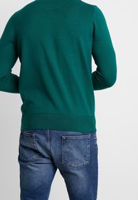 Grüner Strickpullover mit rundem Ausschnitt und geripptem Saum, kombiniert mit blauen Denim-Jeans mit Gesäßtaschen und verwaschener Optik.