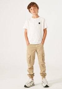 Witte katoenen T-shirt met een zwart logo-embleem, gecombineerd met beige cargobroek met meerdere zakken en elastische boorden, gedragen met witte sneakers.