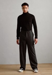 Svart høyhals genser, stripete grå vide bukser og svarte joggesko. Modellen står med avslappede armer på tregulv mot en lys vegg.