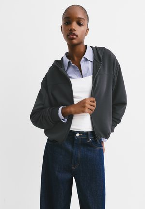 Dark gray zip-up hoodie with a relaxed fit, featuring a collar over a white tank top and a blue-striped shirt underneath. Denim jeans are dark blue.