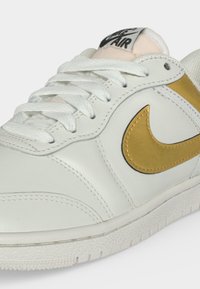 Λευκό δερμάτινο sneaker Nike Air με χρυσό λογότυπο Swoosh, λευκά κορδόνια και υφασμένο πέλμα.