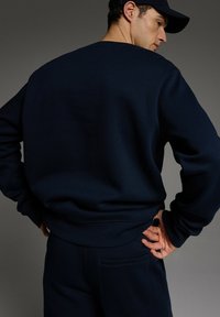 Granatowy sweter z okrągłym dekoltem, elastycznymi mankietami i dolnym wykończeniem oraz teksturowanym materiałem. Charakteryzuje się luźnym krojem, bez widocznych oznaczeń.