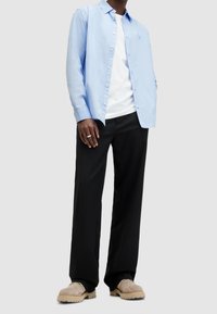 Uomo vestito con camicia azzurro chiaro sopra una maglietta bianca, pantaloni neri e scarpe in suede beige, che sta in piedi contro uno sfondo chiaro e uniforme.