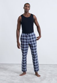 Μαύρο αμάνικο μπλουζάκι και γκρι καρό πιτζαμάνες με μπλε λεπτομέρειες. Οι παντελόνες διαθέτουν ελαστική μέση και κορδόνι. Τα πόδια είναι γυμνά.