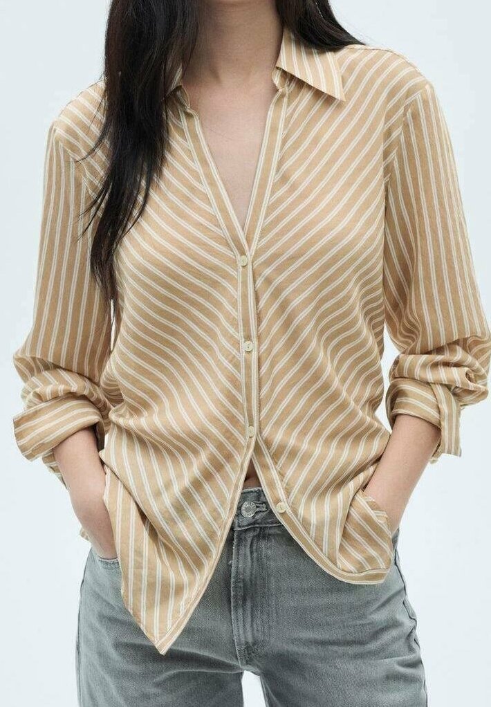 Femme portant une chemise beige et blanche à rayures, manches retroussées, et un jean gris, les mains dans les poches avant.
