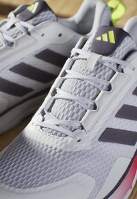 Baskets de sport gris clair avec des rayures grises foncées, tige en maille texturée, lacets blancs et accents roses sur la semelle. Détail au niveau du talon vert.