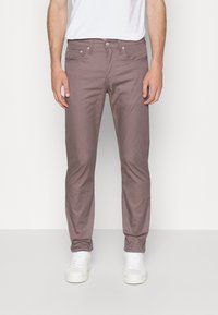 Pantalones chinos de algodón color púrpura claro con un corte slim, bolsillos delanteros y cierre de cremallera, combinados con zapatillas blancas sobre un fondo neutro.