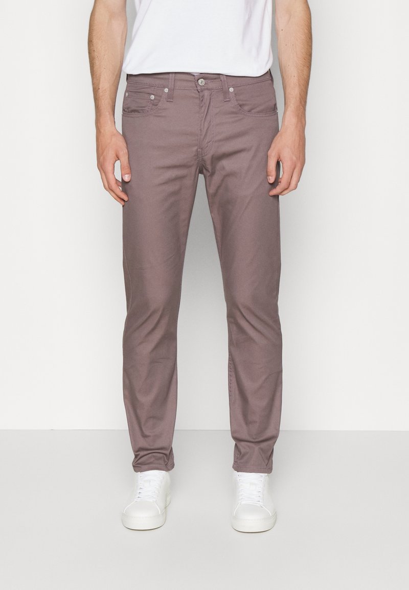 Pantalones chinos de algodón color púrpura claro con un corte slim, bolsillos delanteros y cierre de cremallera, combinados con zapatillas blancas sobre un fondo neutro.