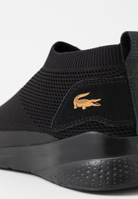 Sneaker nera a maglia con tomaia testurizzata, polsino elastico, suola nera liscia e un piccolo logo di coccodrillo dorato sul lato.